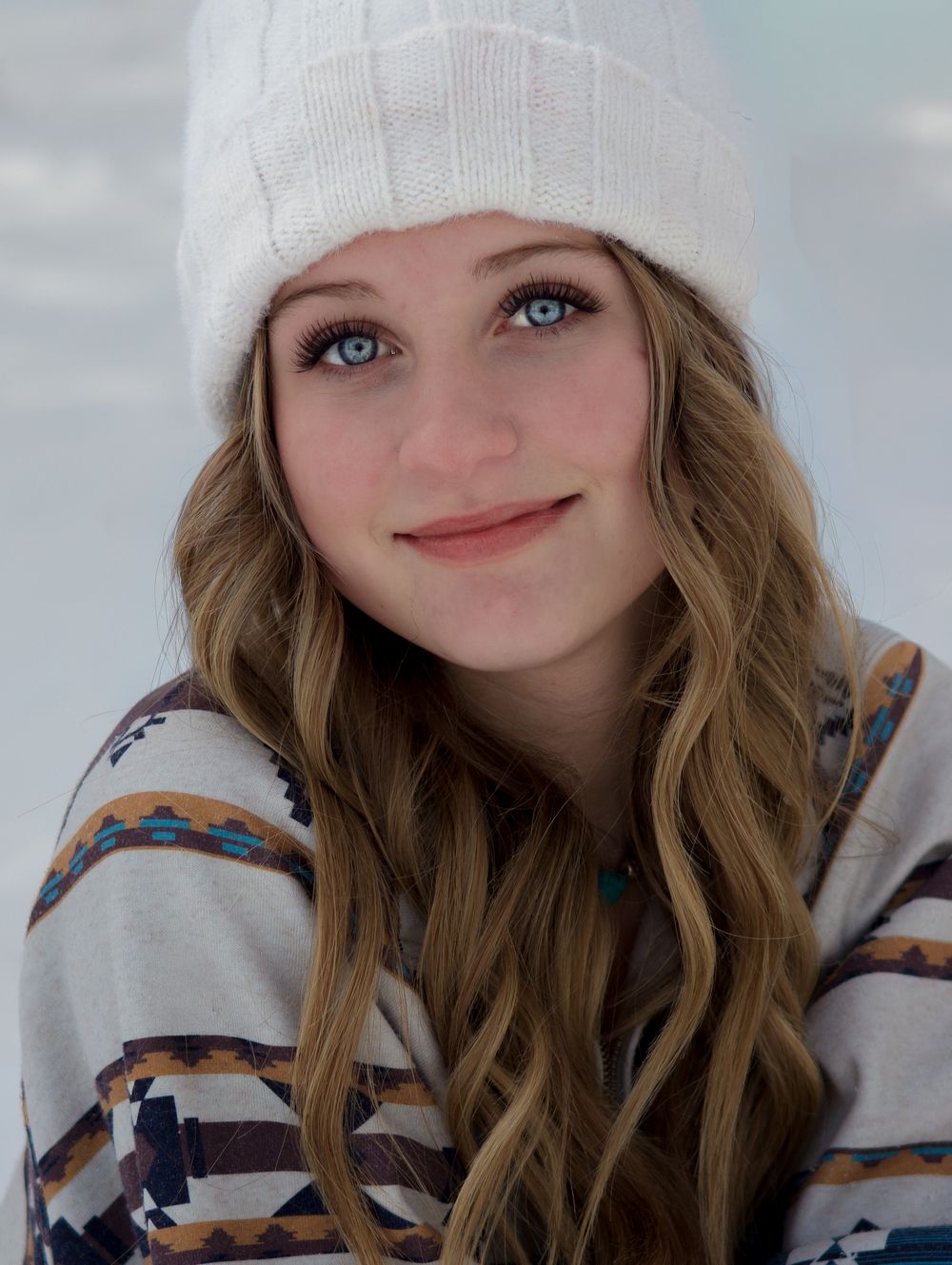 Smiling blue-eyed teen girl in white beanie and Aztec print sweater in winter snow.