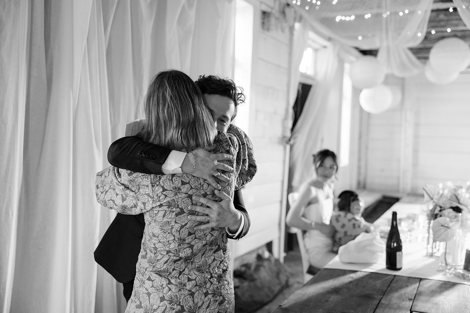 Black and white photo of people embracing at a reception venue decorated with white curtains and paper lanterns.