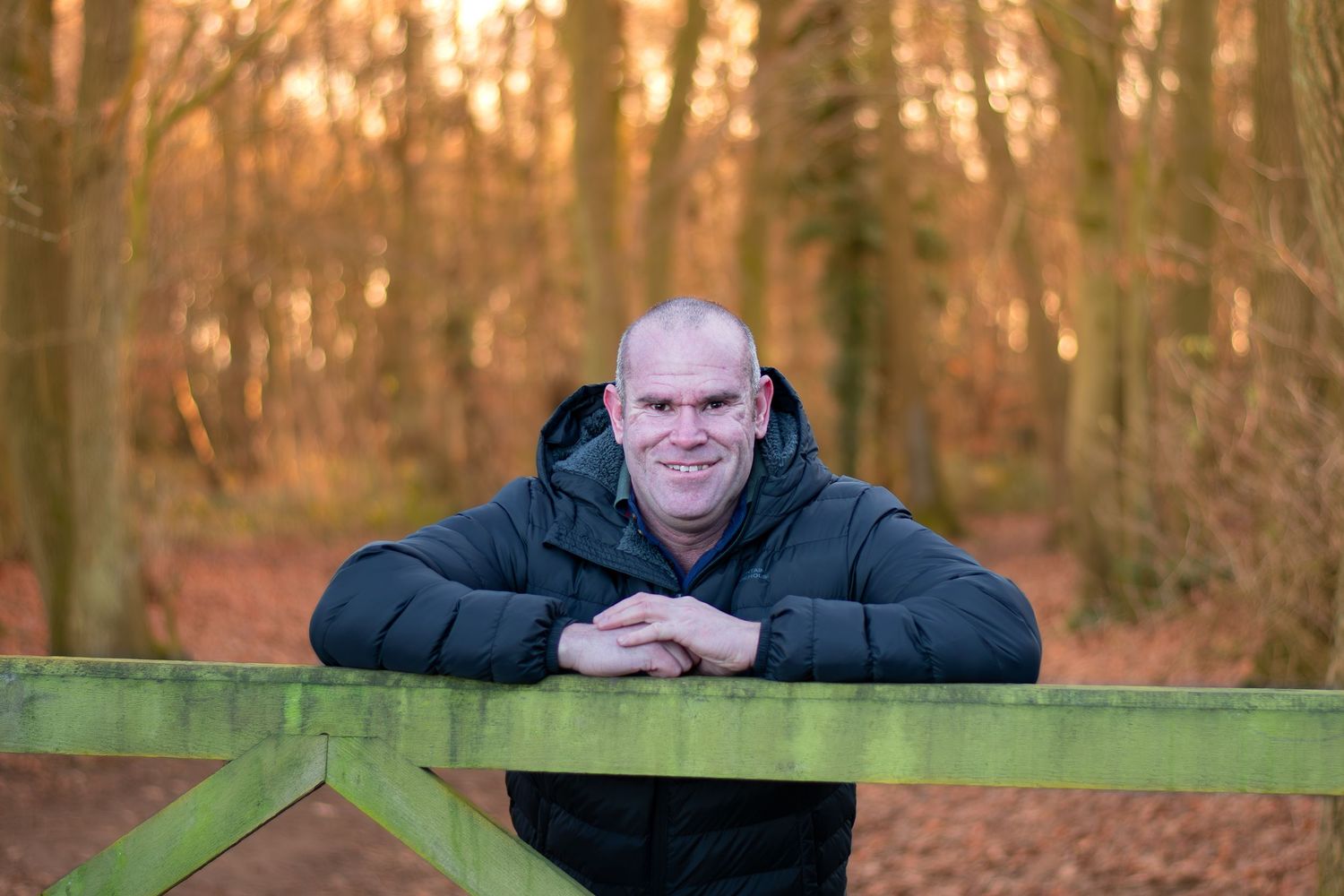Someone in a black winter coat leans on a wooden gate in an autumn forest at sunset in Rotherham, Sheffield, Doncaster South Yorkshire.