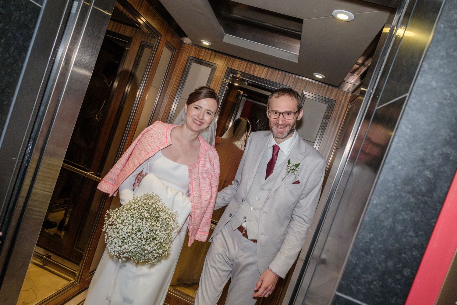A wedding party walks down a hallway in formal attire with bridesmaids in pink dresses and groomsmen in gray suits.