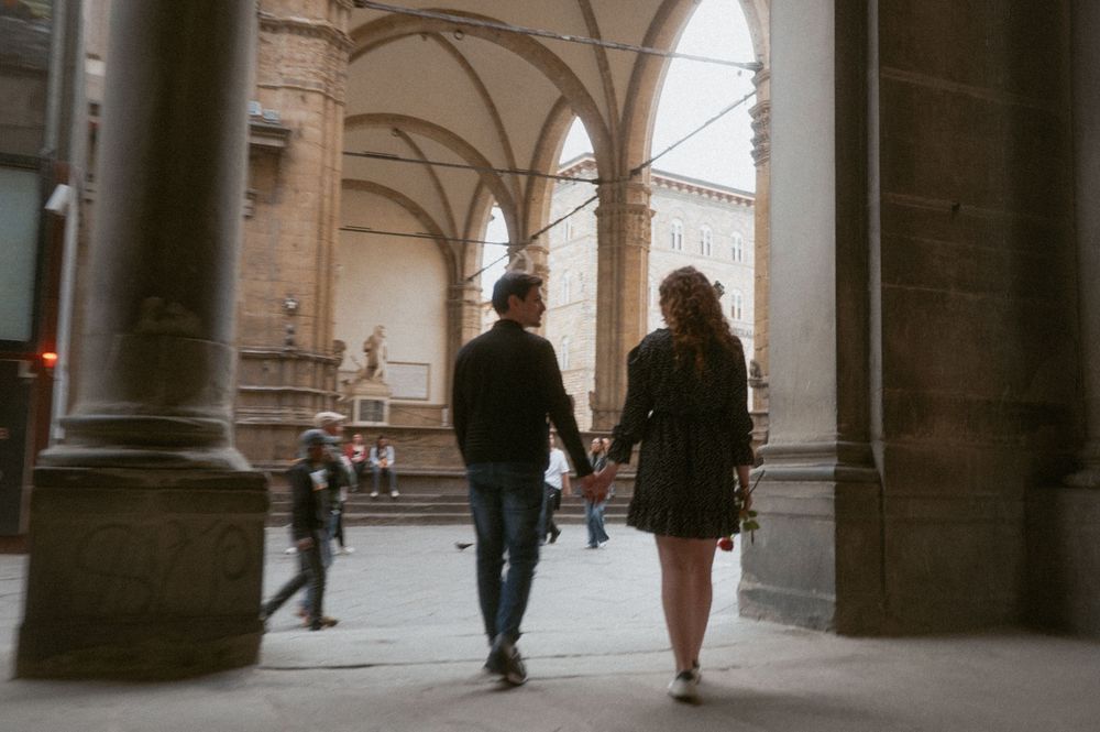 People walking through a historic covered arcade with stone arches and columns in an Italian style architecture.
