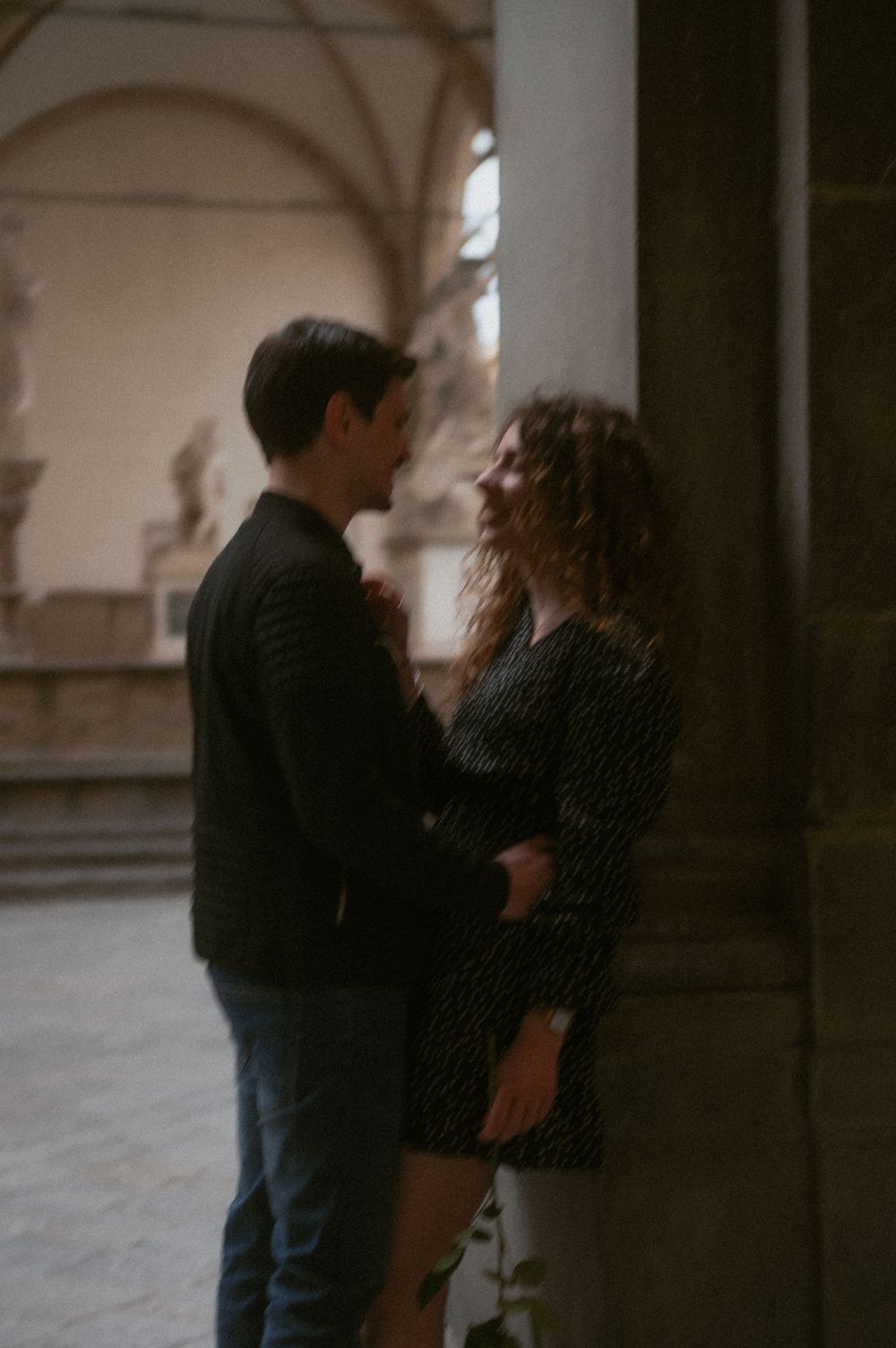 A tender moment between a couple standing in a dimly lit classical colonnade.
