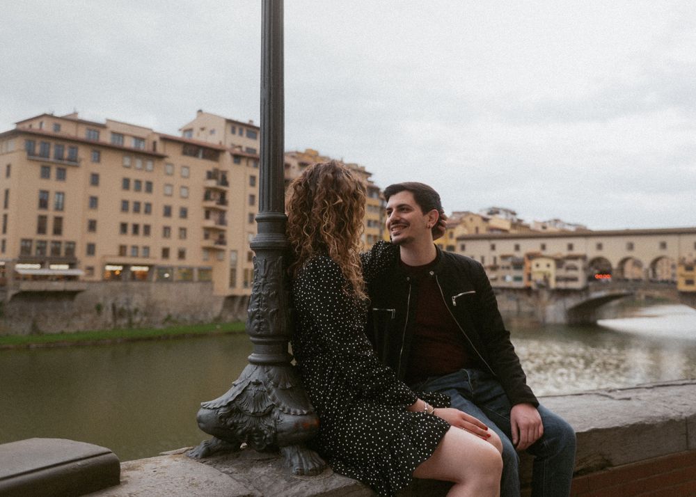 A couple enjoys a tender moment along the Arno River in Florence with city buildings behind them.