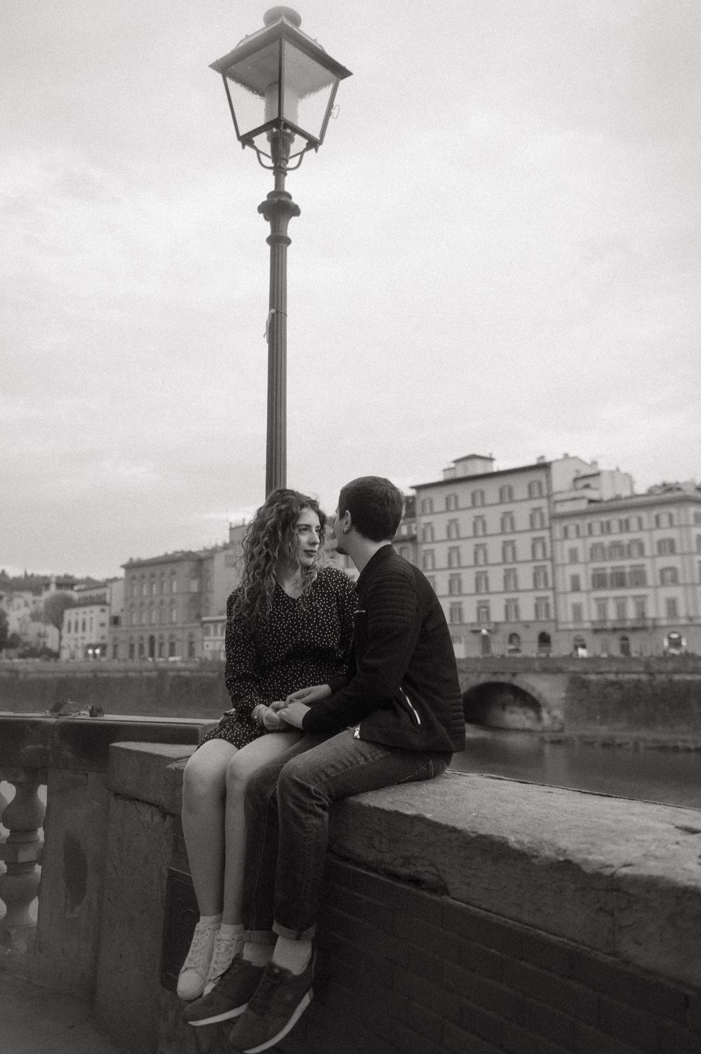Black and white photo of couple sitting on stone wall overlooking Arno River.