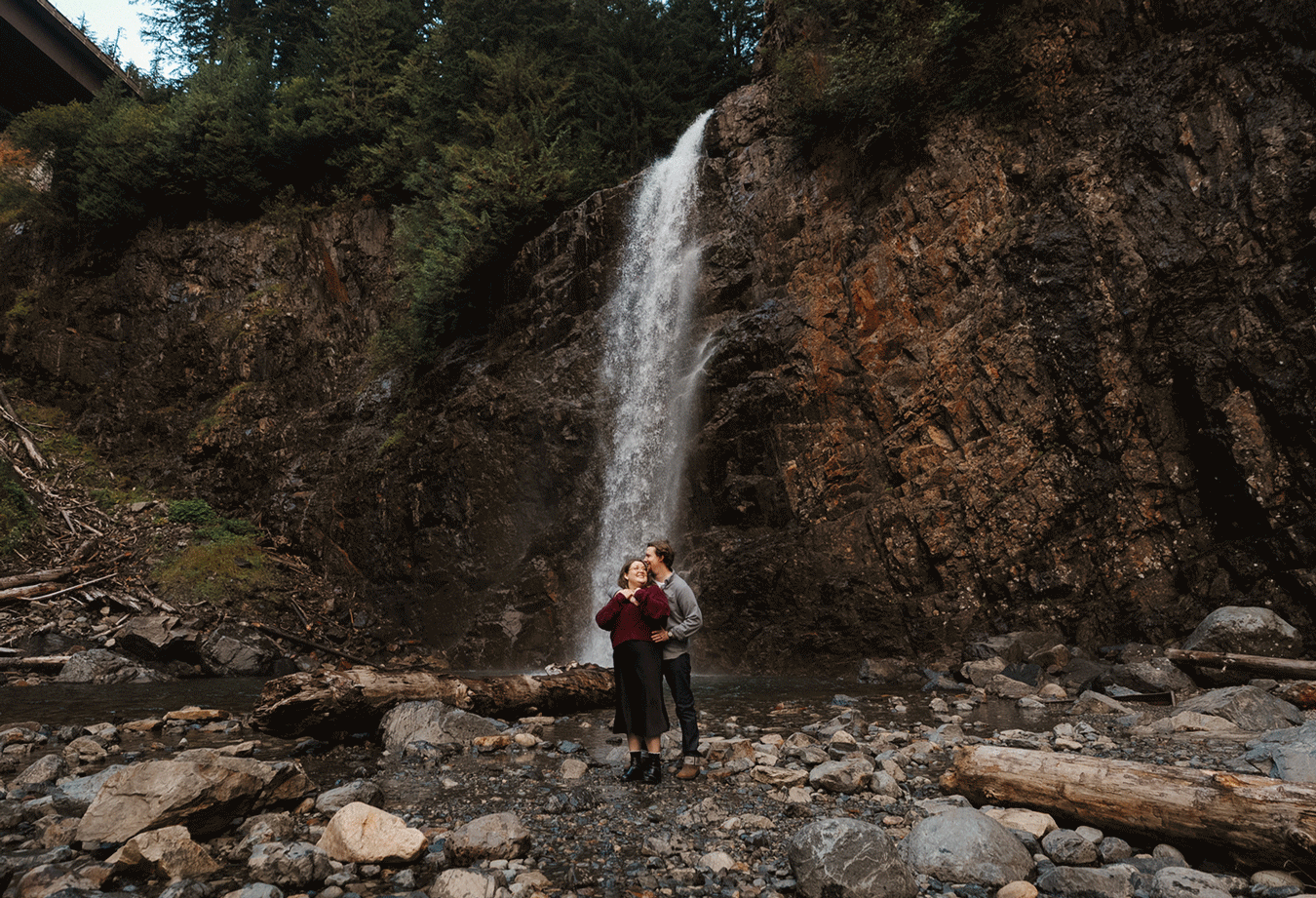 A scenic waterfall cascades down a rocky cliff face in a forest setting with someone standing at its base.