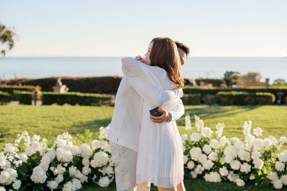 A couple in white outfits embraces during a golden hour photoshoot in a scenic outdoor setting.