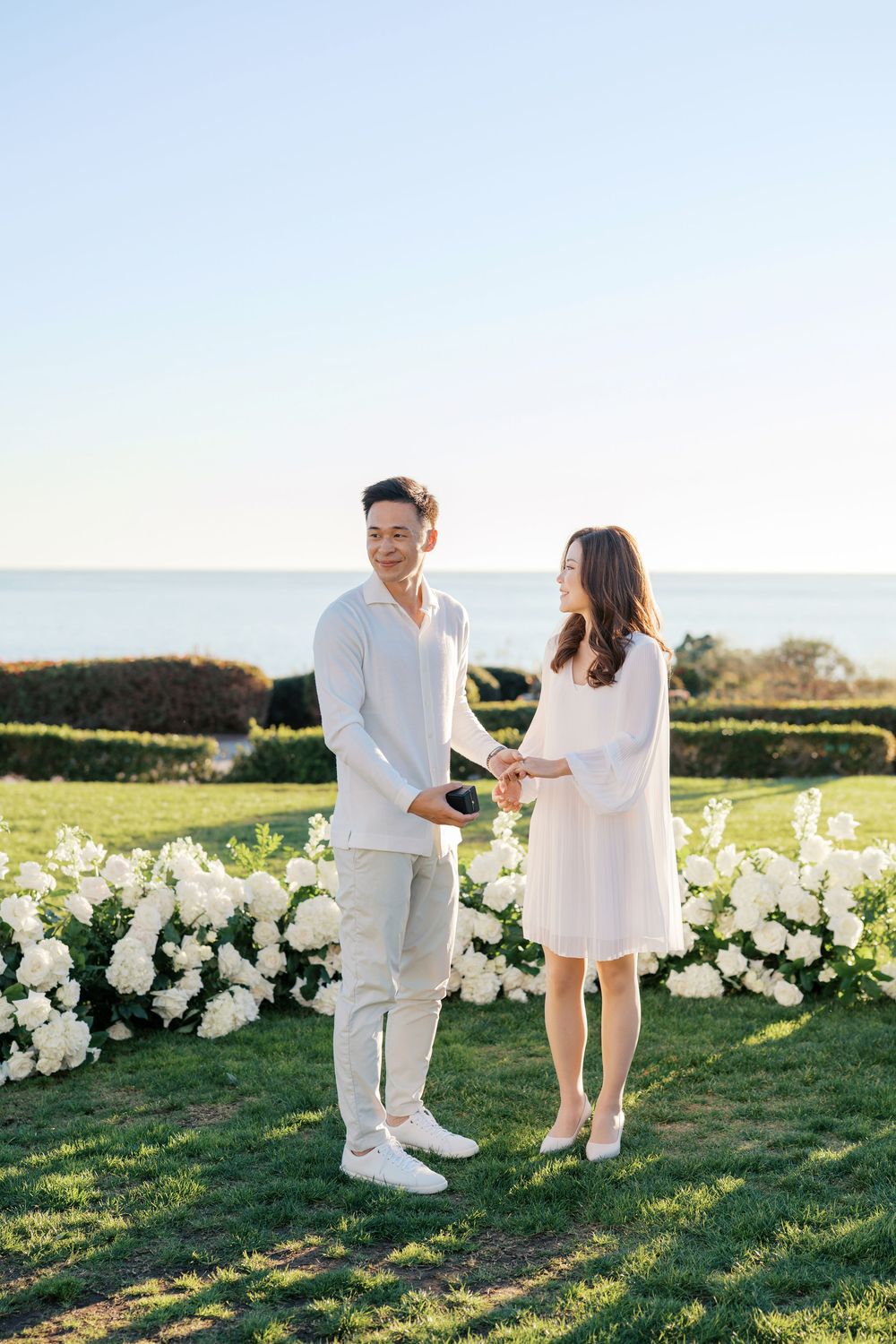 A couple in white outfits dances playfully together on a grassy field with white flowers in the background.