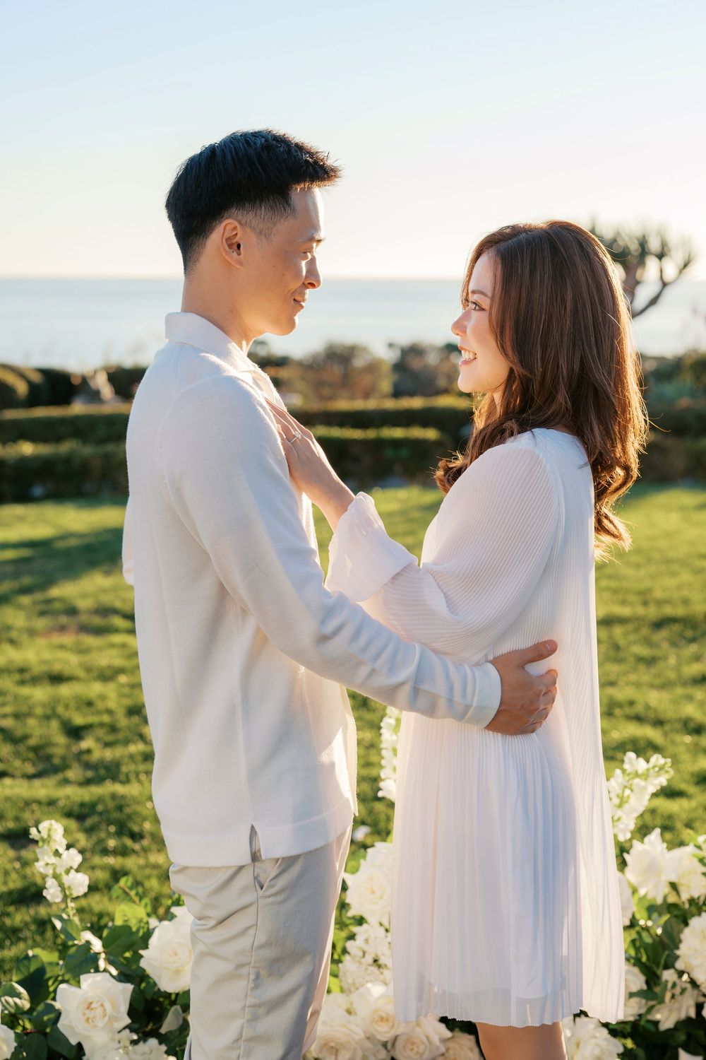 A couple in white outfits embrace in a sunlit field with white flowers in the background.