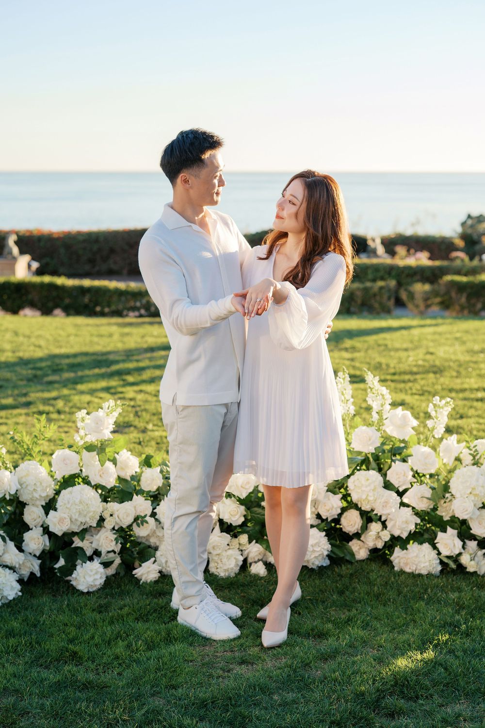 A couple in white outfits dancing together on a grassy hill decorated with white flowers at sunset.