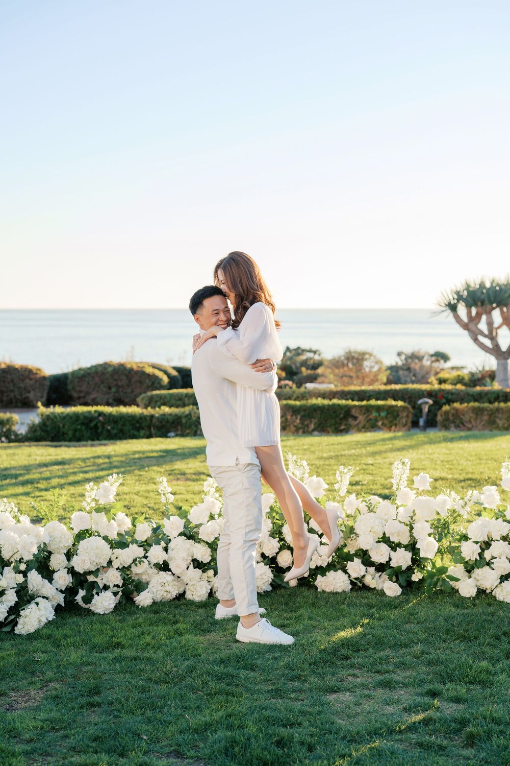 A couple in white outfits embraces on a grassy lawn decorated with white floral arrangements at sunset.