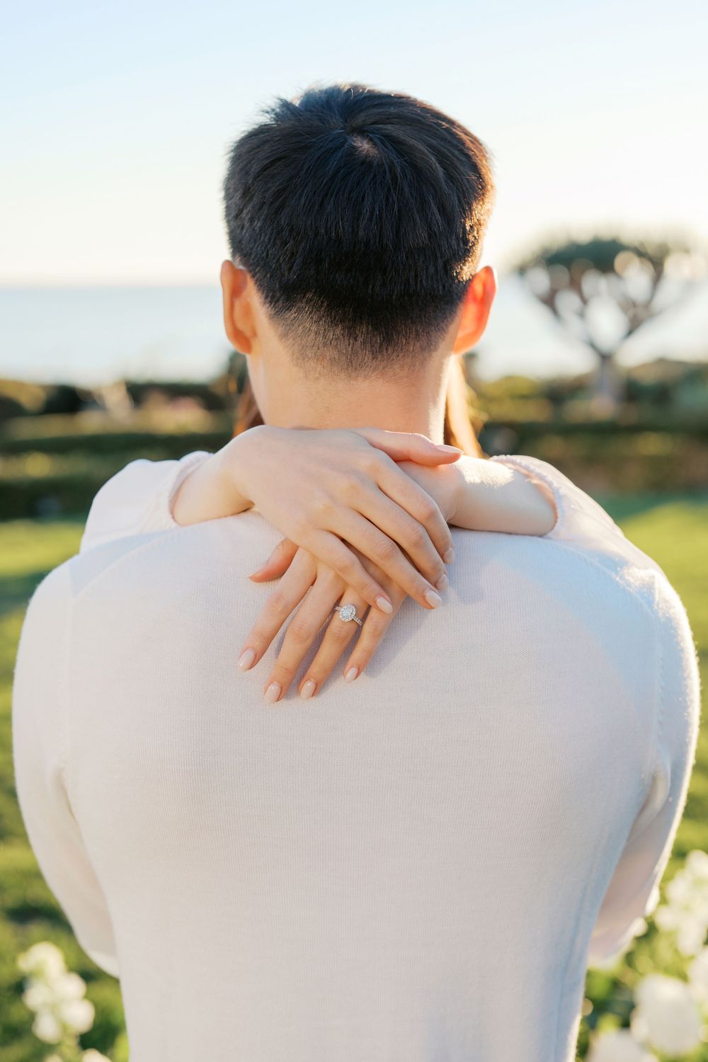 Close-up view of hands tenderly embracing shoulders against a sunlit outdoor backdrop with white flowers.