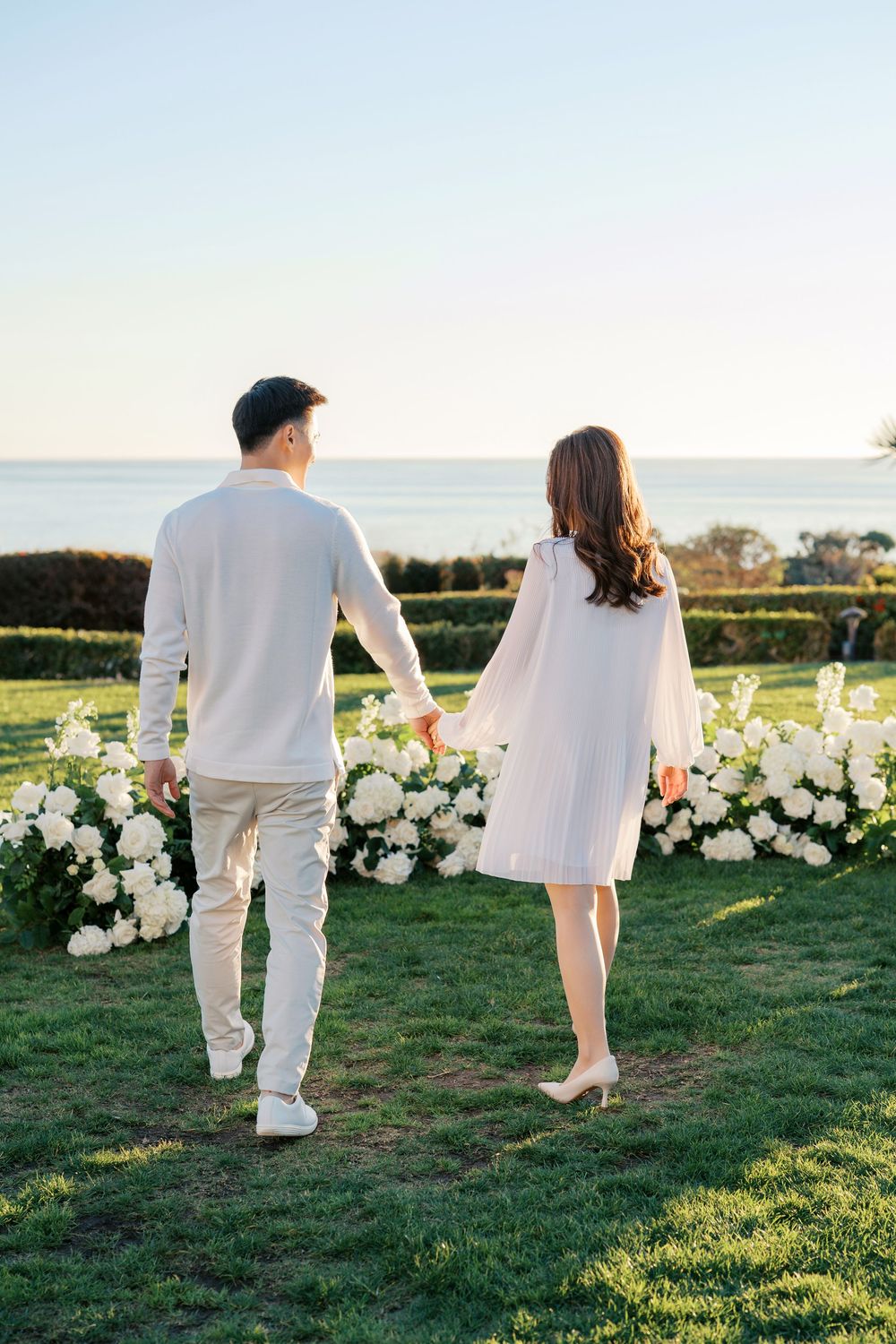 A couple in white outfits walks together on a grassy lawn decorated with white floral arrangements.
