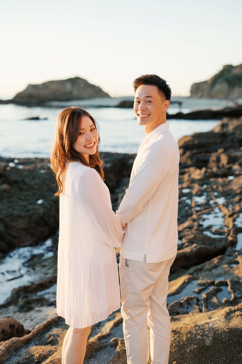 A couple in white outfits stands together on a rocky beach at sunset, creating romantic silhouettes against the coastline.