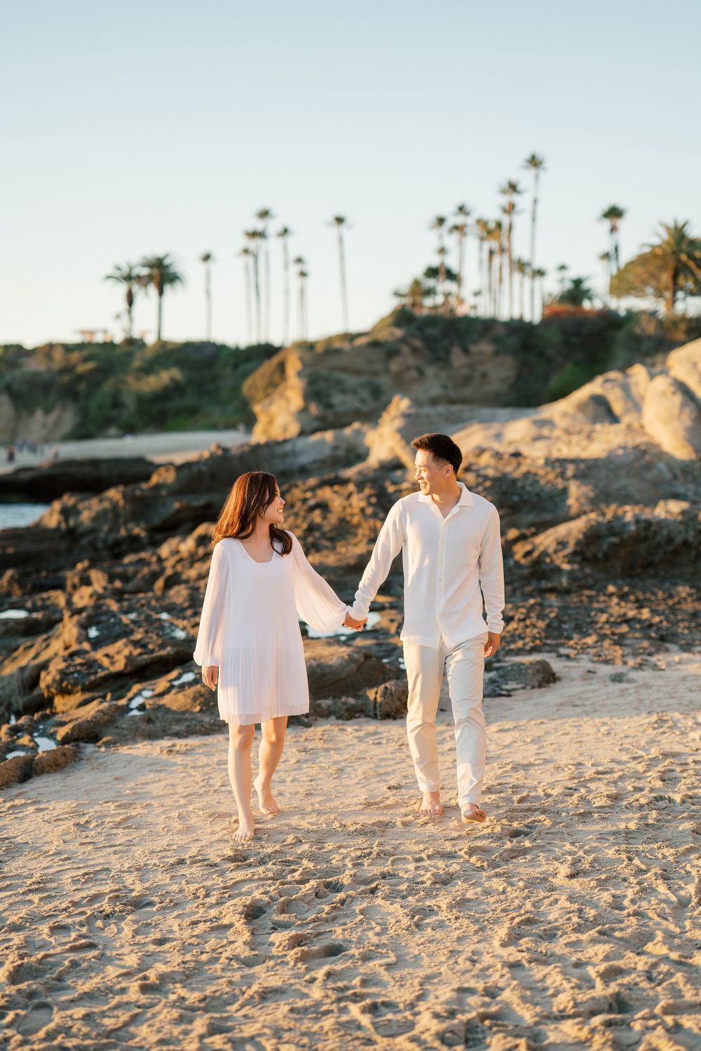 A couple in matching white outfits strolls together along a sandy beach with palm trees in the background.