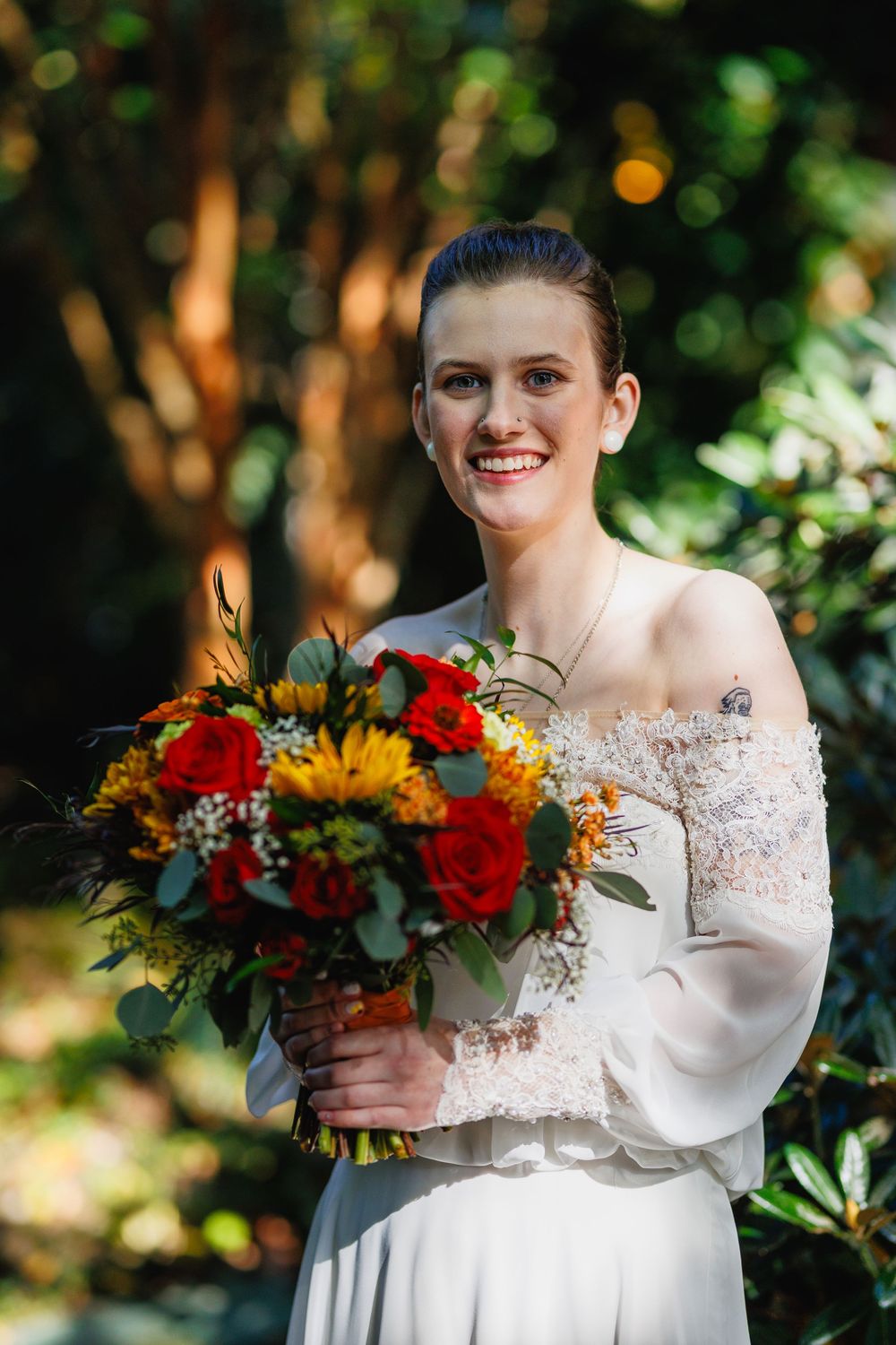 Smiling bride in white off-shoulder gown holds a vibrant fall bouquet of sunflowers and red roses outdoors.