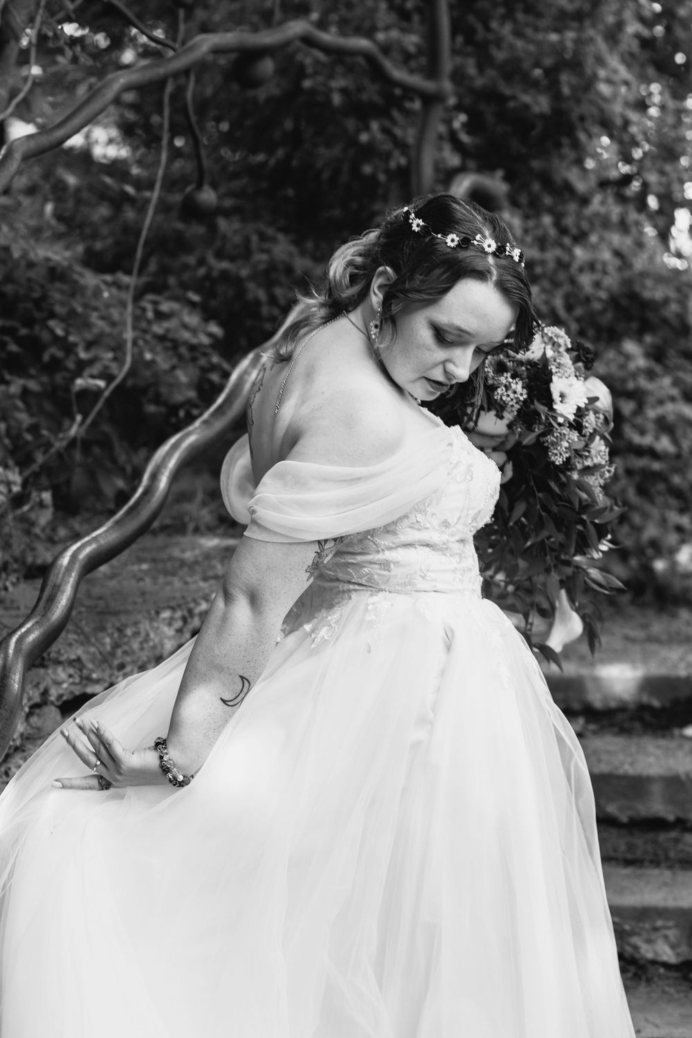Black and white photo of a bride in an off-shoulder gown holding a floral bouquet outdoors.