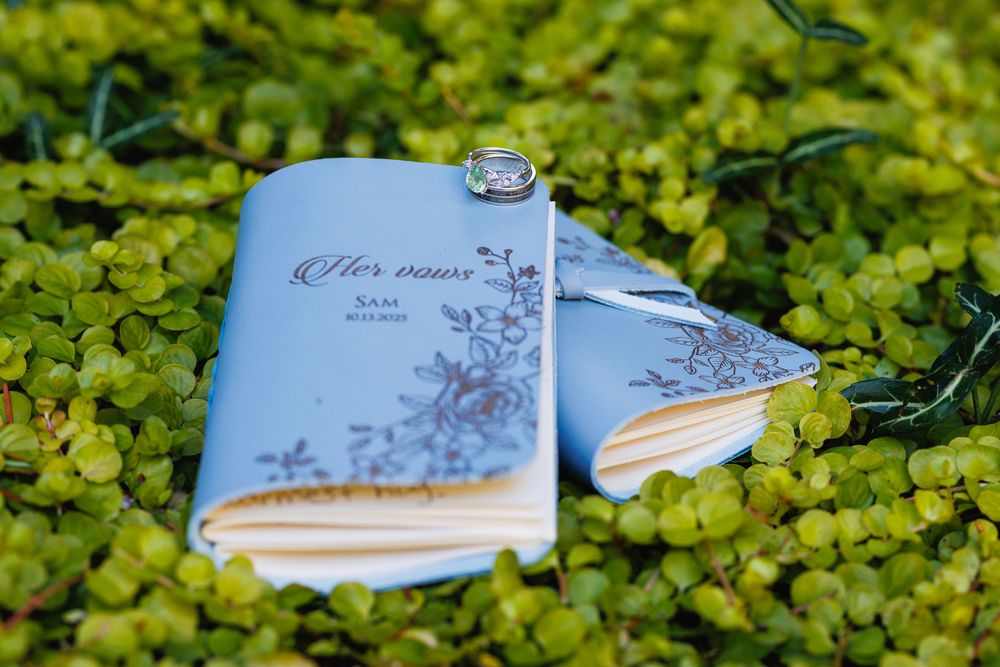 Light blue wedding vow books with floral design and a diamond ring resting on green foliage.