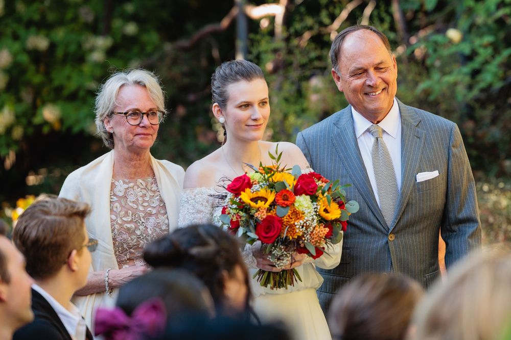 Bride walks down the aisle with parents holding a vibrant sunflower bouquet at outdoor wedding.
