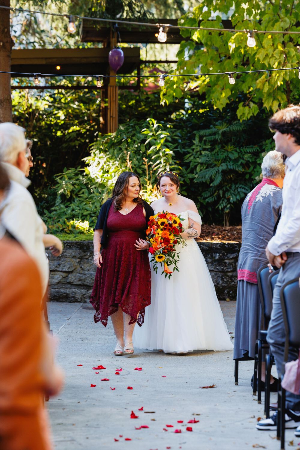 Bride in white gown walks the aisle with a woman in burgundy dress holding sunflower bouquet.