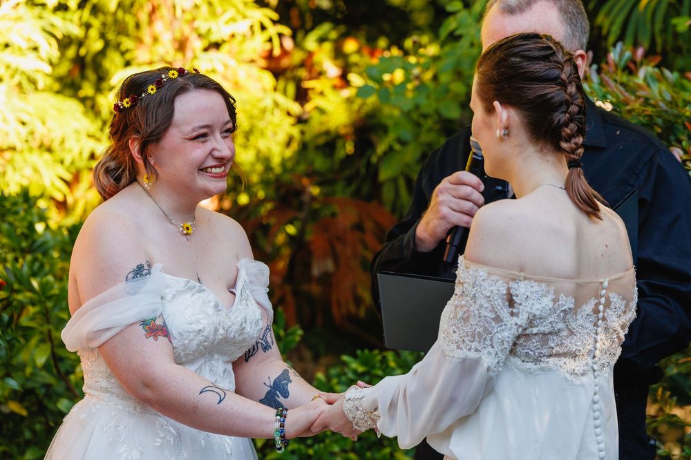 Two brides in white gowns hold hands smiling during outdoor garden wedding ceremony.