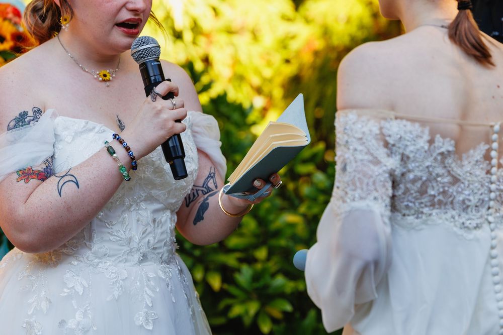 Bride with tattoos reading vows from a book while holding a microphone at outdoor wedding.