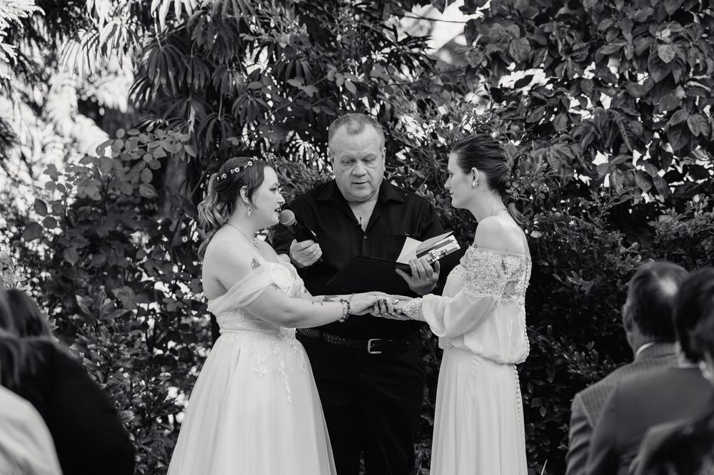 Two brides share their first kiss surrounded by rose petals at an outdoor garden wedding.