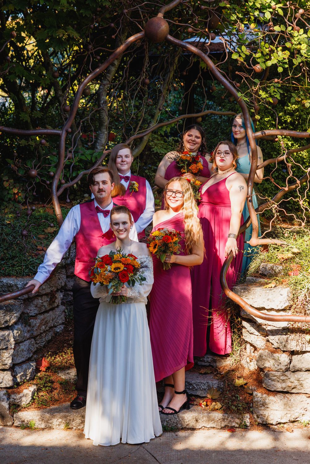 Wedding party in magenta dresses and burgundy vests poses under a rustic arch surrounded by autumn foliage.