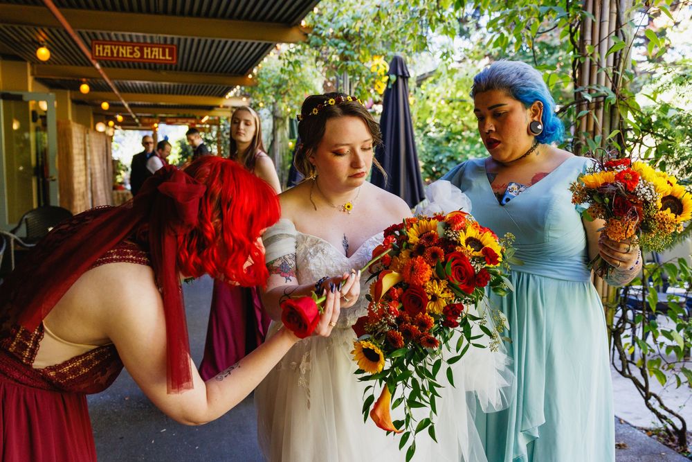 Bride receives her colorful sunflower and fall flower bouquet from a bridesmaid with red hair outdoors.
