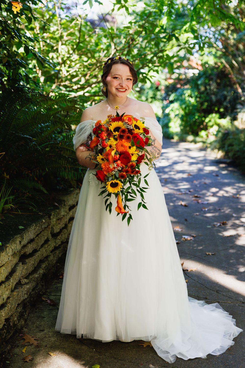 Bride in off-shoulder white ball gown smiles holding a cascading sunflower and red rose fall bouquet outdoors.
