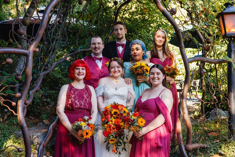 Wedding party in magenta and teal attire poses together under a twisted arch in a lush garden setting.