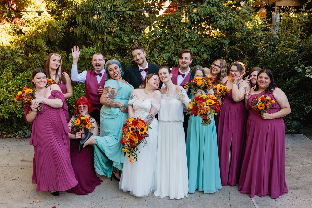 Large joyful wedding party with two brides and attendants in magenta and teal dresses celebrate outdoors.
