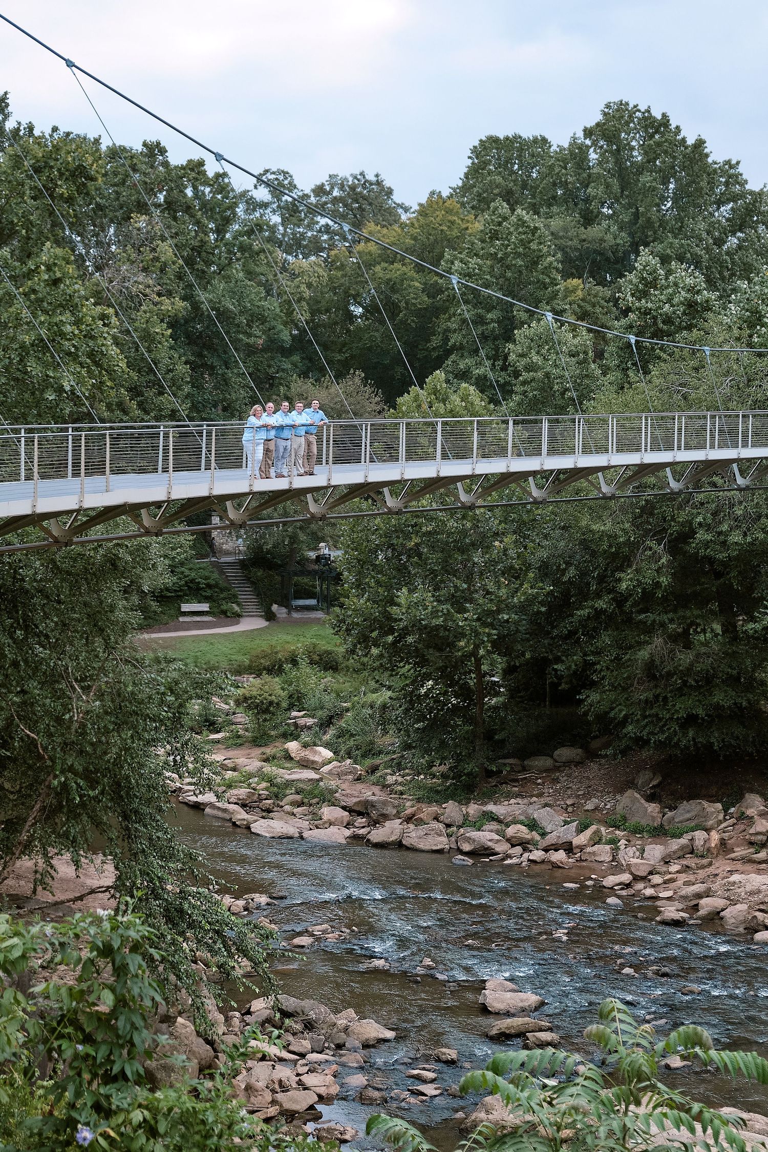 A suspension footbridge spans over a rocky creek with flowing water, surrounded by lush green trees on both sides.