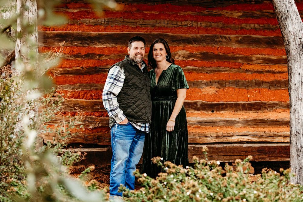 A couple stands together against a rustic red log cabin wall surrounded by autumn foliage.
