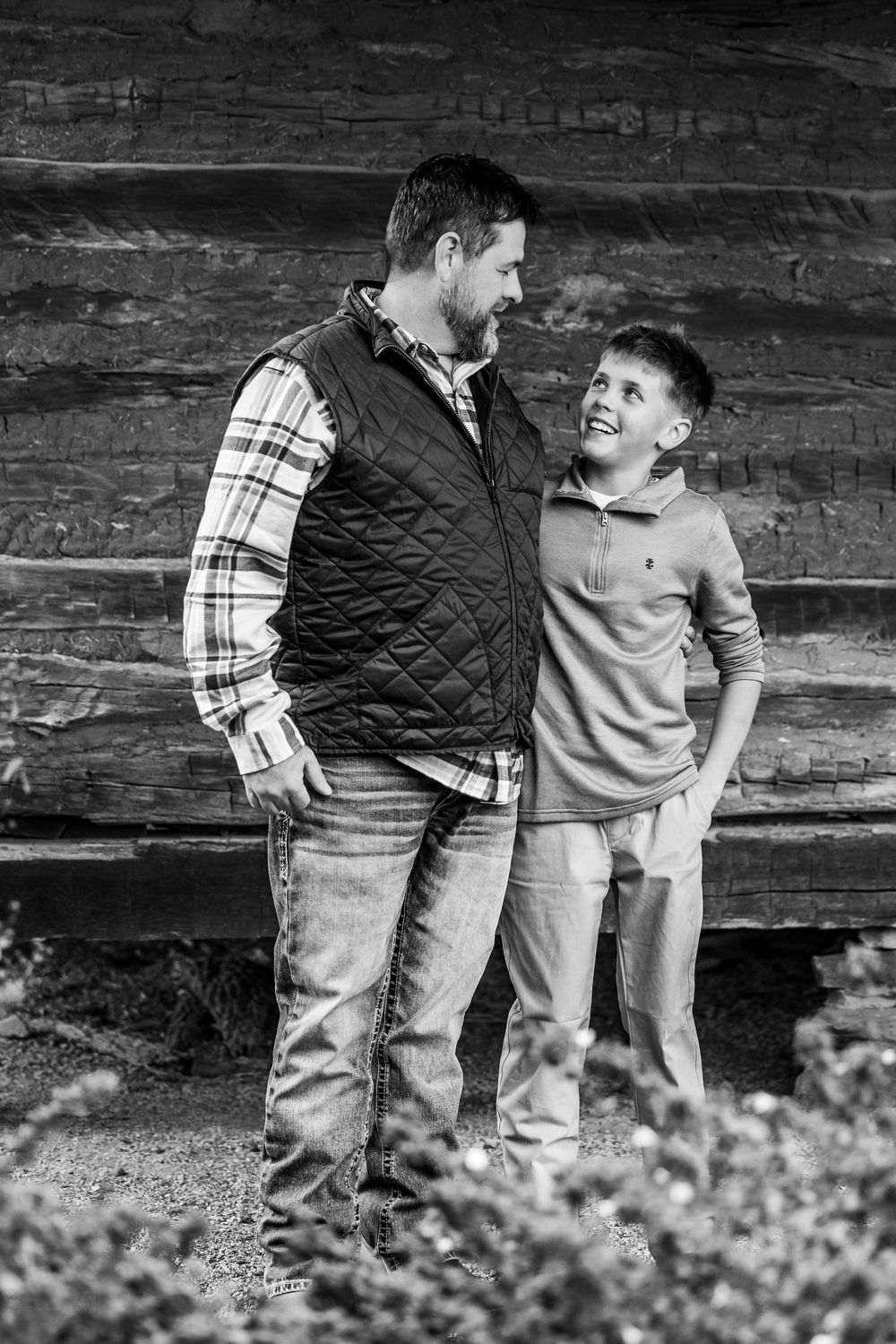Black and white photo showing two people in casual clothing leaning against a rustic wooden wall outdoors.