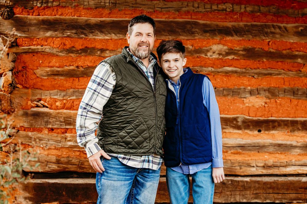 Two people stand smiling together against a rustic wooden log cabin wall wearing casual vests and plaid shirts.