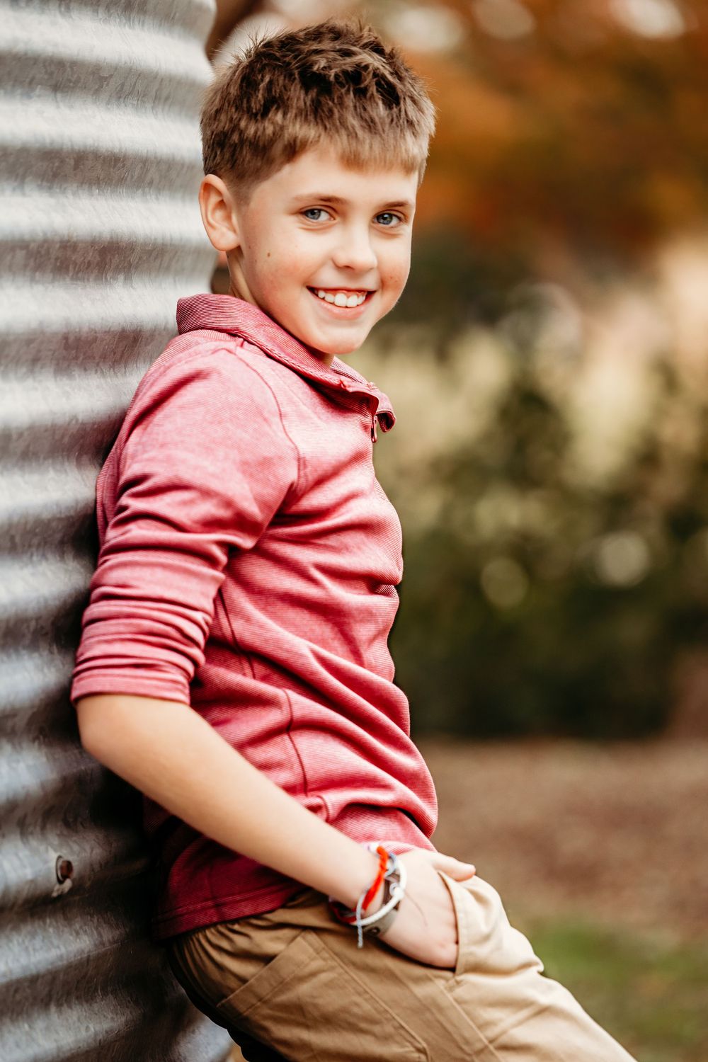 A child in a coral pink collared shirt and khaki pants sits casually outdoors against a blurred background.