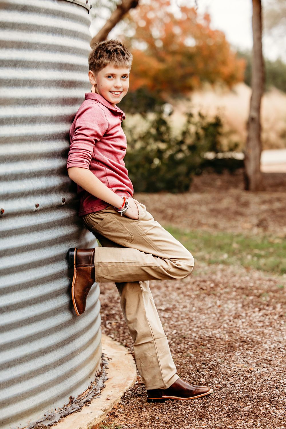 A child in a red polo shirt and khaki pants sits casually on a curved metal surface outdoors in autumn.