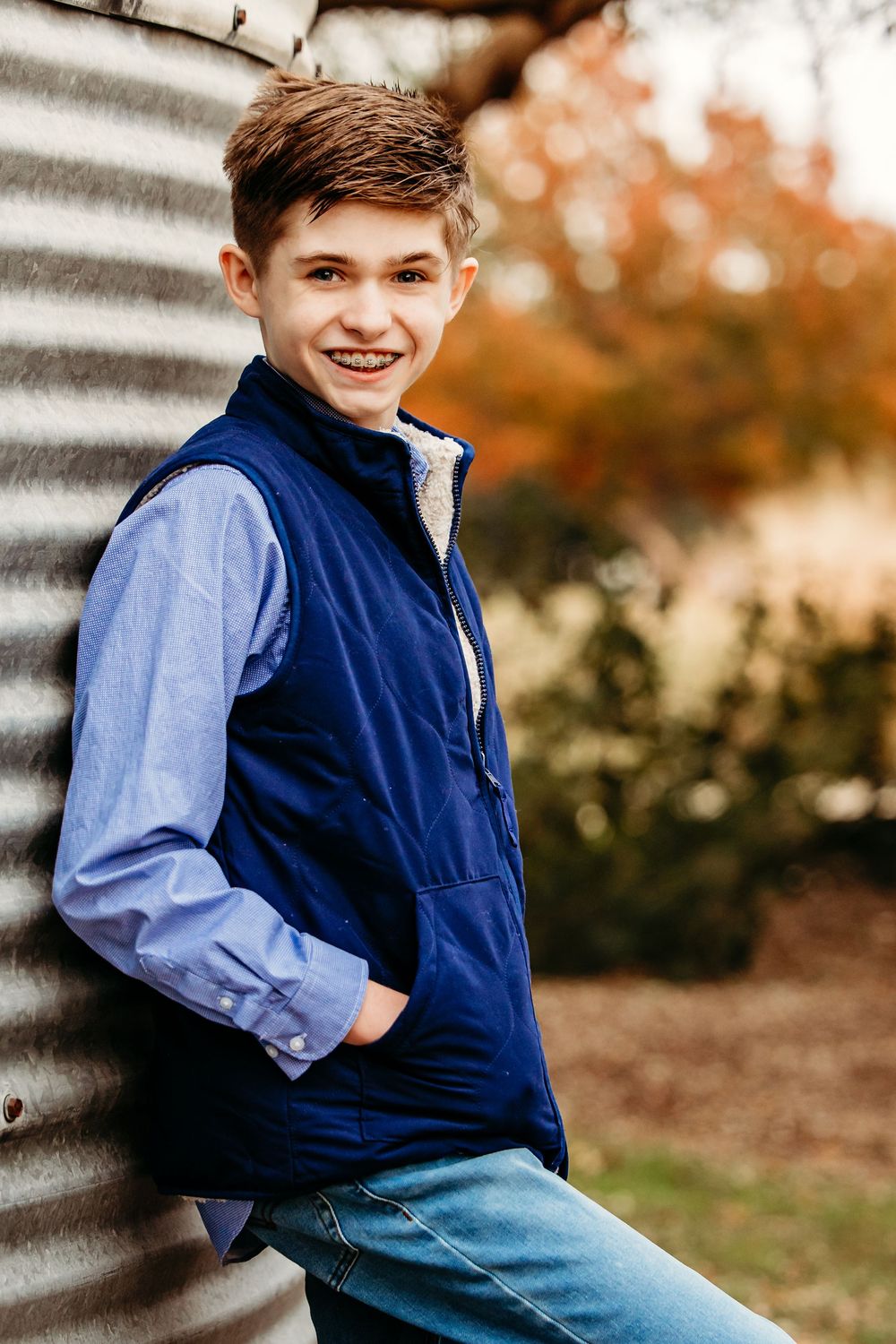A youth in a navy fleece vest and blue button-down shirt poses outdoors against an autumn background.