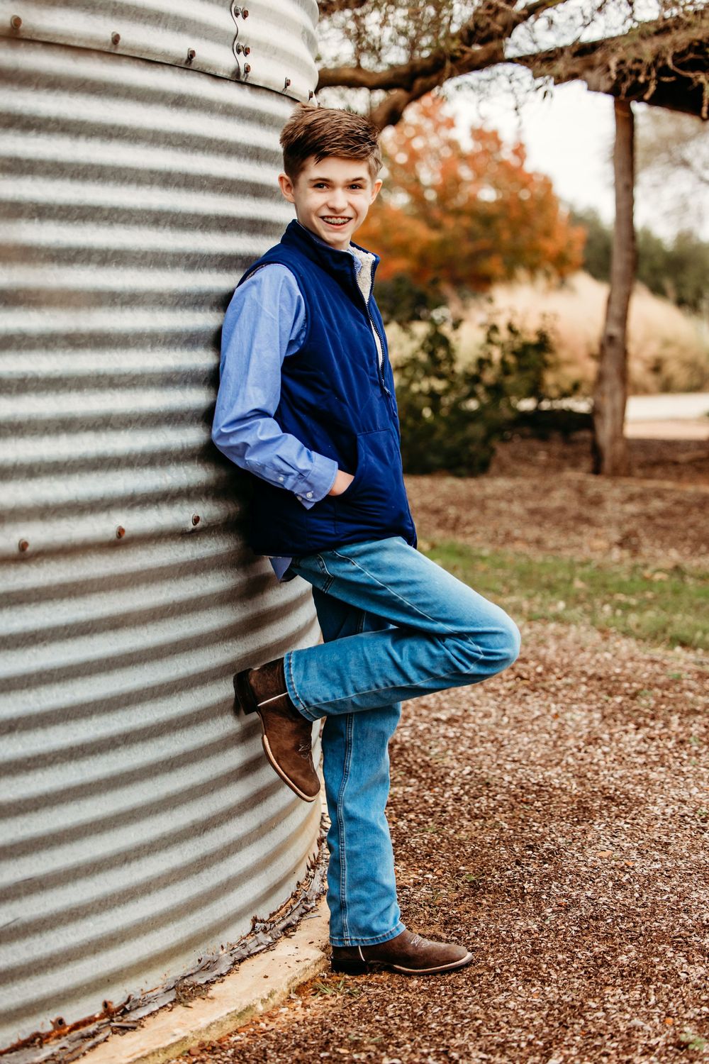 A person in a blue vest and jeans poses casually against a corrugated metal grain silo wall in an autumn setting.