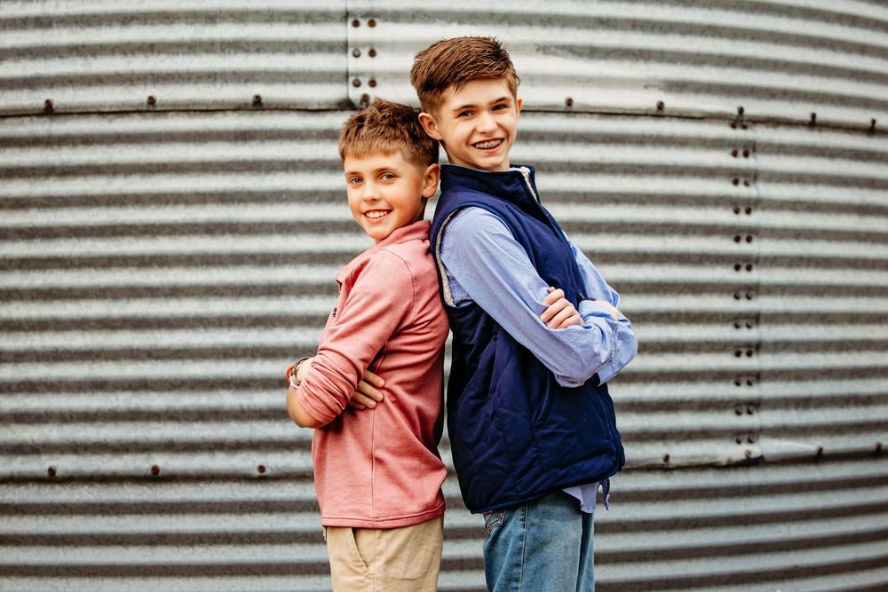 Two young boys stand back to back wearing casual vests against a corrugated metal wall.
