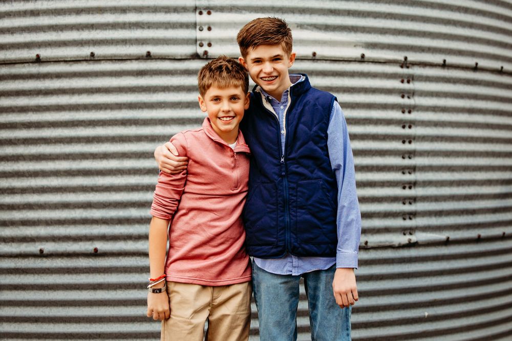Two young boys in casual clothing pose and smile together against a striped metal wall background.