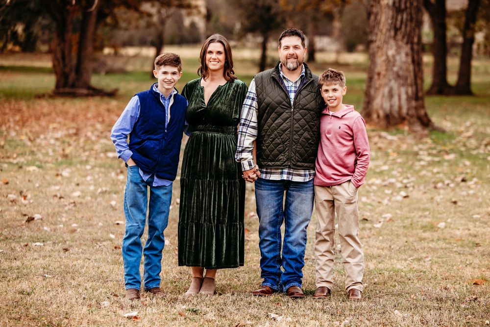 A family stands together in a wooded park setting during autumn with fallen leaves scattered on the ground.