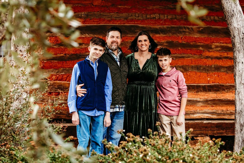 Family poses together for outdoor autumn portraits in front of a rustic red wooden barn wall.