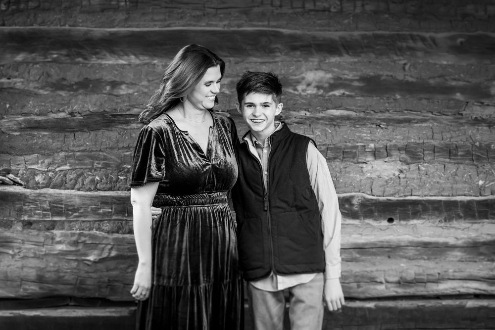Black and white portrait of a parent and child standing together against a rustic wooden wall background.