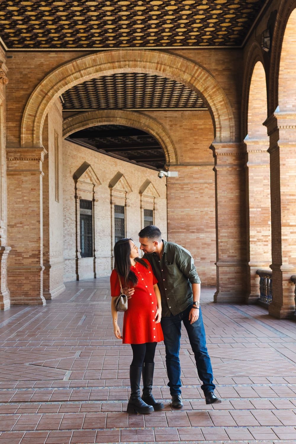 A couple stands together under ornate arches with brick columns and decorative ceilings in a historic building.