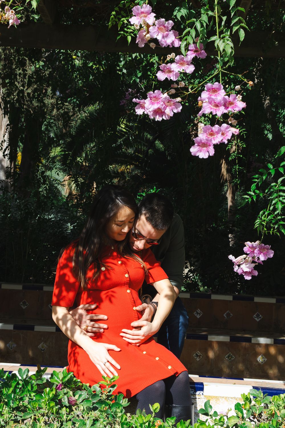 A couple in a garden embraces tenderly under pink flowering bushes and green foliage on a sunny day.