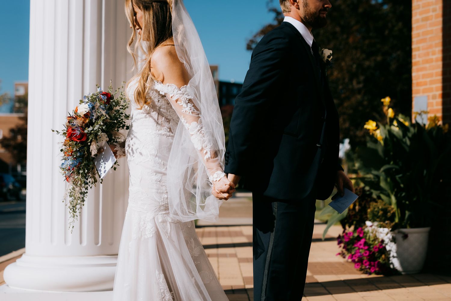 Intimate first touch portrait as bride and groom hold hands facing away from each other surrounded by columns outside a Catholic church