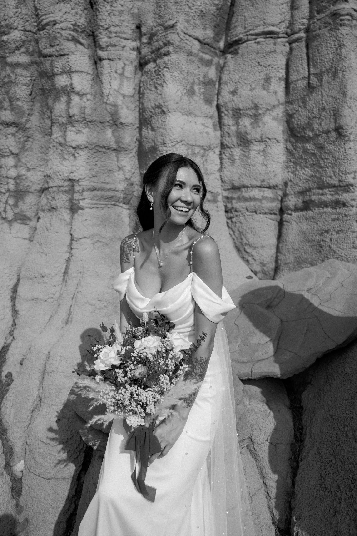A bride in an off-shoulder white dress holds a colorful bouquet while posing against dramatic rock formations.