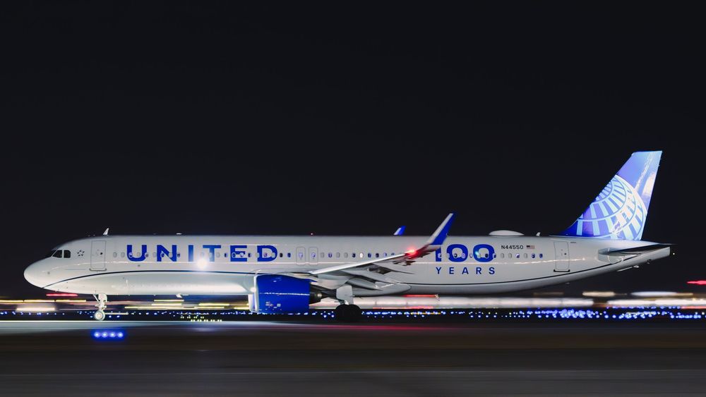 United Airlines 757 aircraft with 100 Years livery on tarmac at night, illuminated by airport lights.