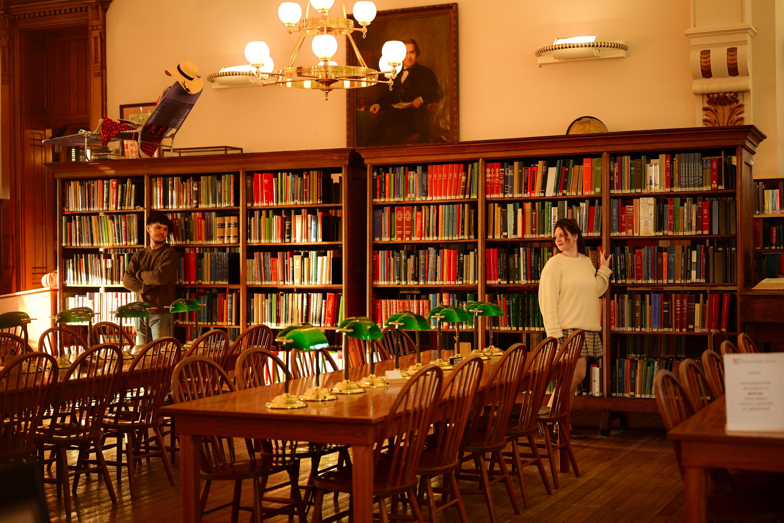 A warm, cozy library interior with wooden tables and chairs illuminated by soft lighting and lined with bookshelves.